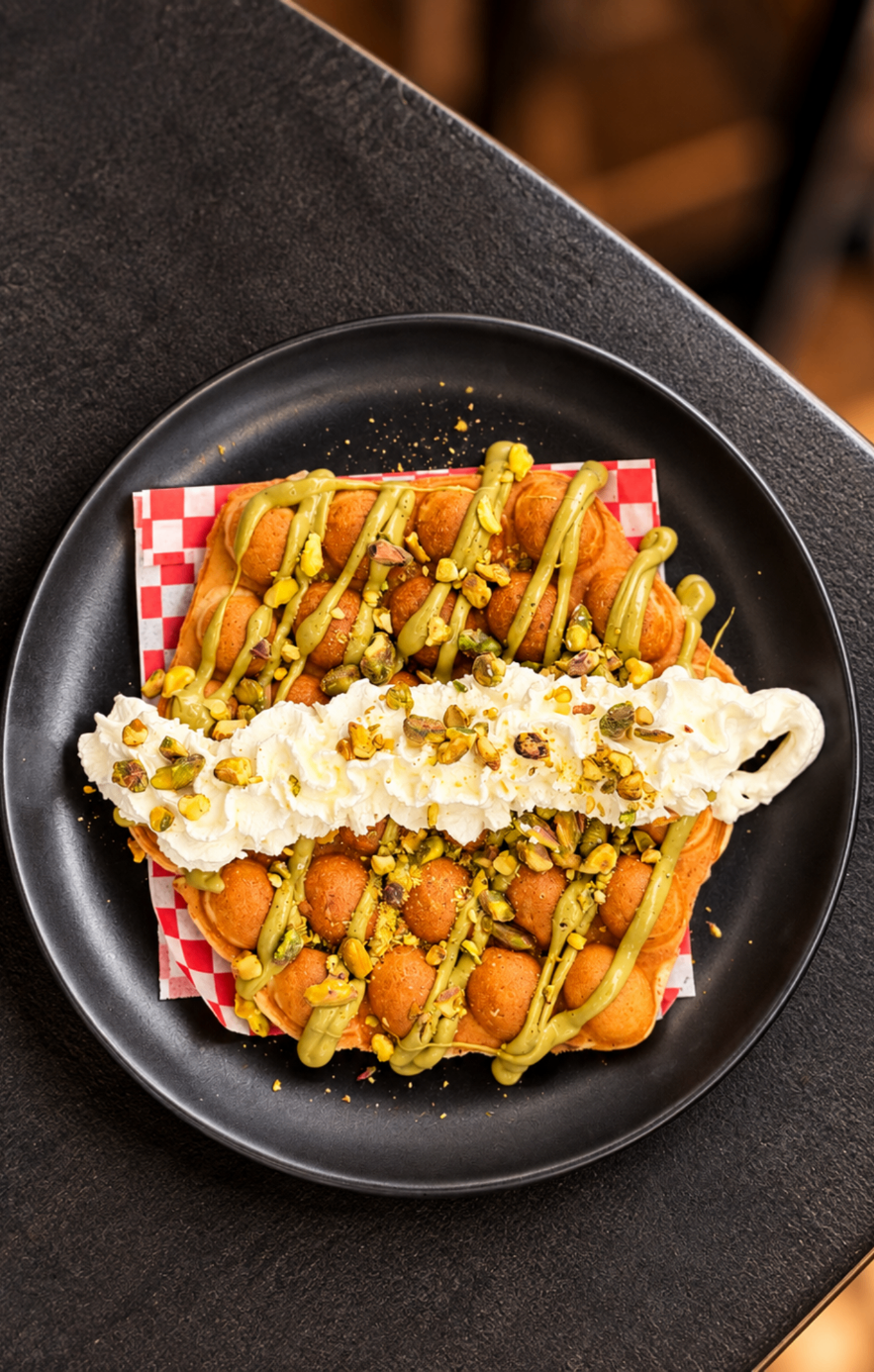Bubble waffle garnie de sauce pistache, chantilly maison et pistaches concassées, dans une assiette noire