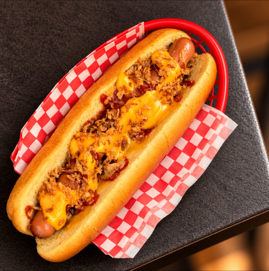 Hot-dog nappé de cheddar fondu et d'oignons frits, dans un panier rouge sur papier damier rouge et blanc